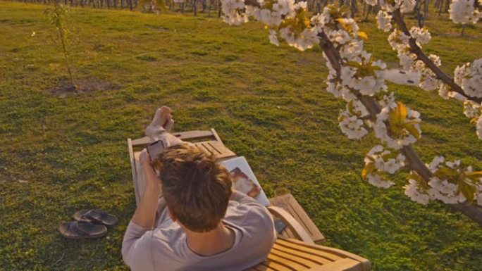 带着智能手机的曼女士在阳光明媚的田园诗般的山坡上的躺椅上放松。这个人正在躺椅上休息。