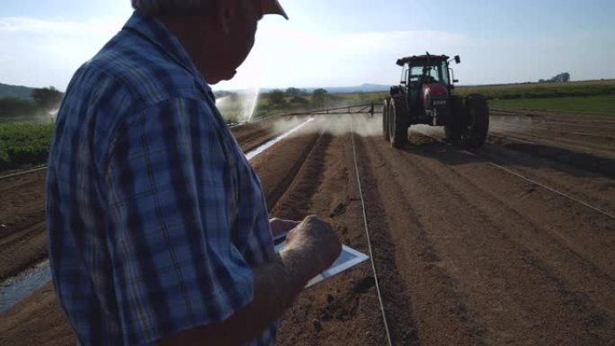 使用数字平板电脑的农民的4k裁剪视图，监视拖拉机在大型蔬菜农场上用农药喷洒新耕作的土壤