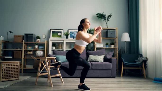 运动的年轻女士蹲在家里的一条腿上独自在公寓里锻炼