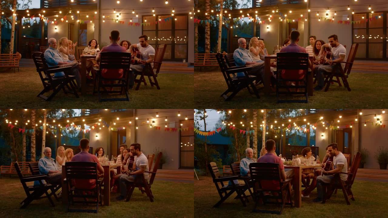 坐在餐桌上的英俊年轻人为朋友弹吉他。家人和朋友在夏季晚上的花园聚会庆典上听音乐。