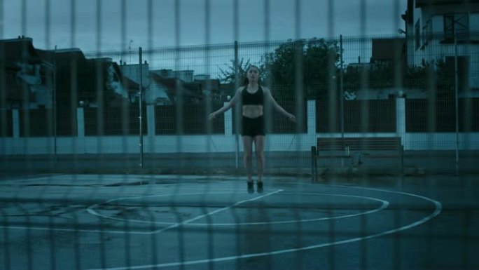 穿着黑色运动上衣和短裤的美丽精力充沛的健身女孩正在跳绳。她正在一个有围栏的室外篮球场里锻炼身体。从栅
