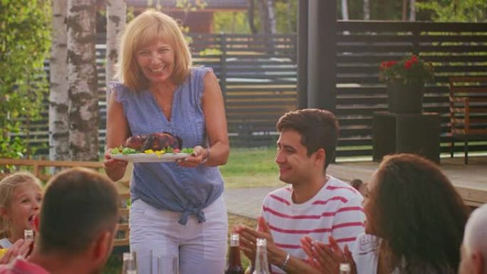 在家庭花园聚会上，母亲将烤鸟的菜带到餐桌上。家人和朋友聚集在大桌子旁。吃喝玩乐。