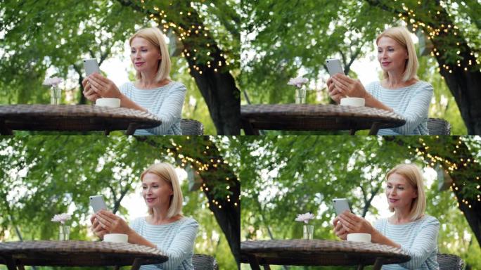 迷人的成熟金发女郎使用智能手机在露天咖啡馆享受夏日
