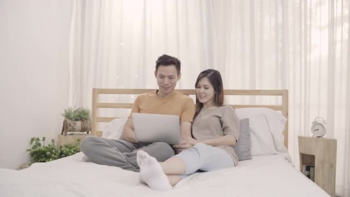 亚洲夫妇在卧室放松时躺在床上使用笔记本电脑，夫妇在家里用电脑玩社交媒体。夫妇在家里使用放松时间的概念