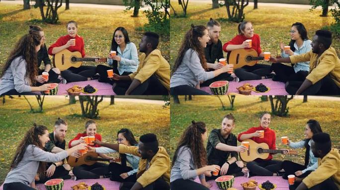 开朗的年轻人多种族群体在秋天野餐时，用饮料碰杯，然后坐在草地上的毯子上喝吉他和食物。