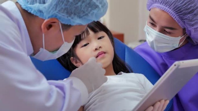 友好的医生和女婴在牙医办公室看平板电脑的肖像。可爱的小女孩坐在牙科椅上接受牙科治疗。医学，口腔医学和