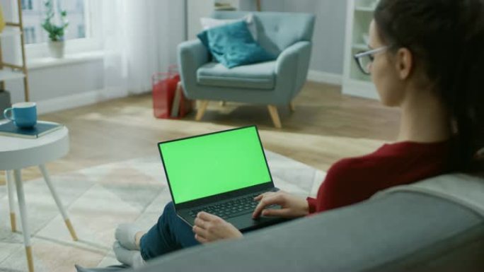 家里的年轻女子在带有绿色模拟屏幕的笔记本电脑上工作。她坐在他舒适客厅的沙发上。肩膀上的相机拍摄