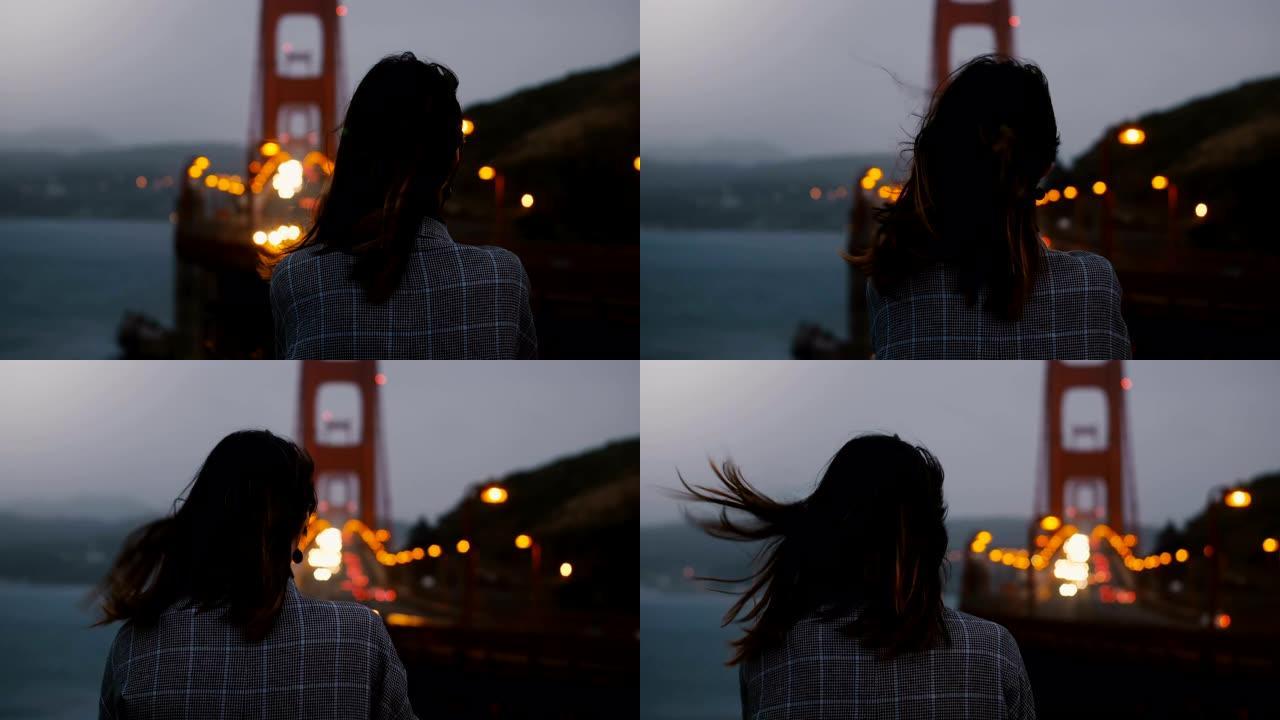 傍晚时分，年轻女子独自站在强风下观看标志性的金门大桥交通景观。
