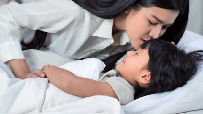 美丽的年轻亚洲母亲亲吻她的婴儿睡在床上。母亲带着生病的孩子在家。母亲正在给生病的女儿量体温。亚洲小女