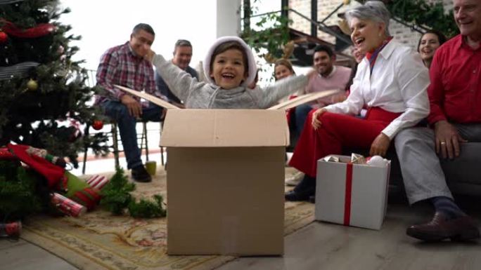 可爱的小男孩作为圣诞礼物从盒子里出来，家人在家里的背景下兴奋地鼓掌