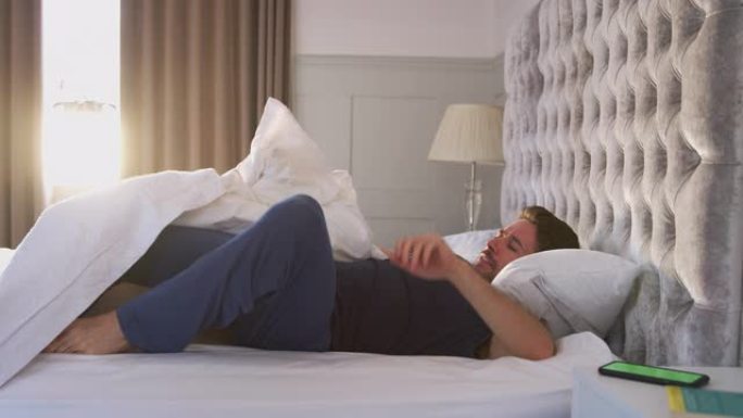 在床上醒来的男子伸手在伸展和起床之前关闭手机上的警报
