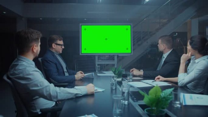 深夜在公司会议室: 董事会，高管和商人坐在谈判桌旁，交谈并使用绿色模拟屏幕墙电视进行视频电话会议。