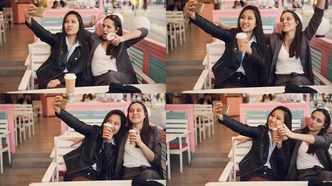 无忧无虑的女孩正在自拍，一起坐在咖啡馆里喝点饮料，并使用智能手机。年轻女性正在摆姿势，碰杯并展示手势