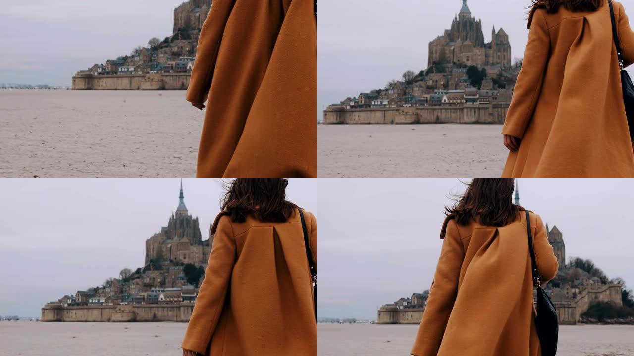 穿着优雅外套的特写快乐旅游女人走向诺曼底迷人的圣米歇尔山城堡慢动作。