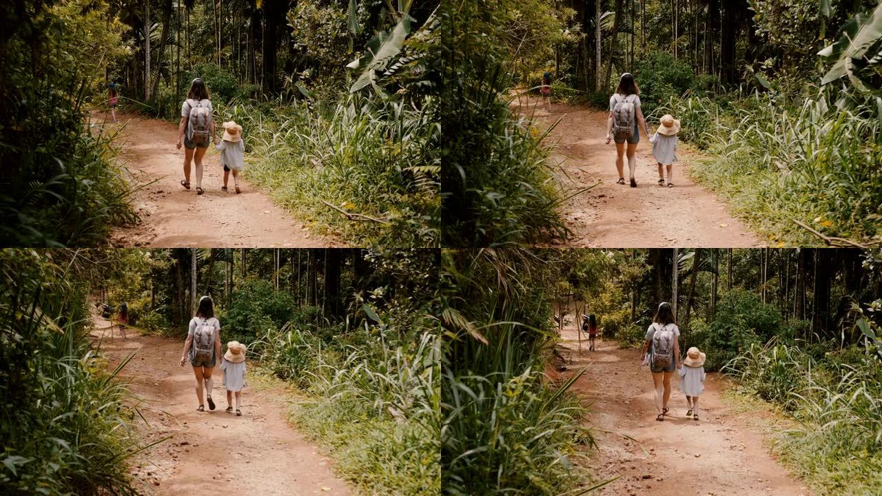 后视图快乐的年轻母亲和戴着大草帽的小孩一起走在陡峭的森林道路上慢动作。