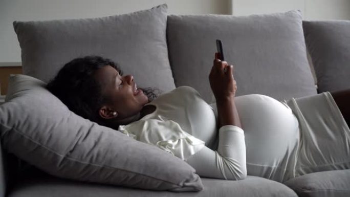 漂亮的黑人孕妇躺在沙发上放松，看着智能手机上的社交媒体