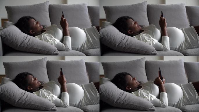 漂亮的黑人孕妇躺在沙发上放松，看着智能手机上的社交媒体