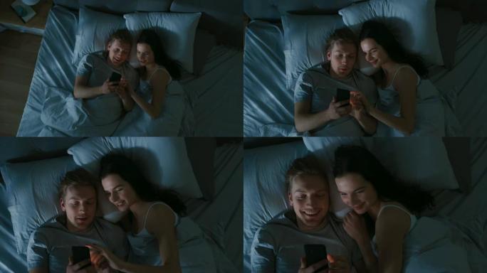 幸福的年轻夫妇晚上在床上使用智能手机，他们共享屏幕，浏览社交网络，共享图片，使用互联网并获得乐趣。用