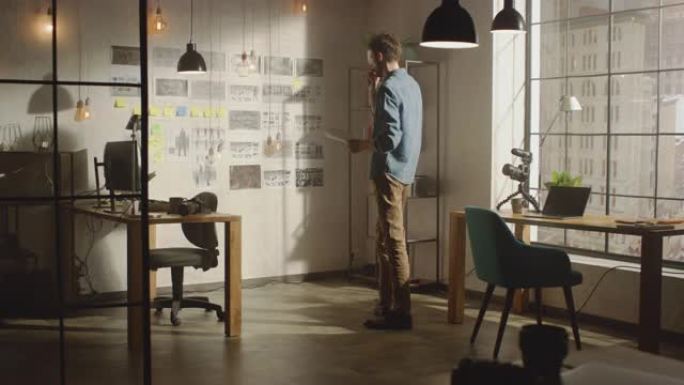 年轻的有创造力的人站在他凉爽的阁楼办公室里，带着有组织的情绪板站在墙旁边。他在一张纸上做笔记。窗外阳