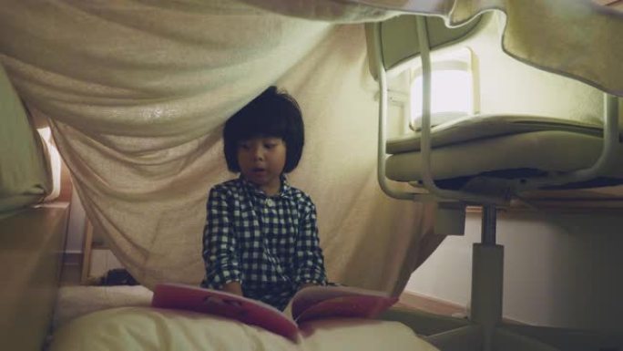 亚洲男孩在毯子堡垒下用手电筒阅读童话
