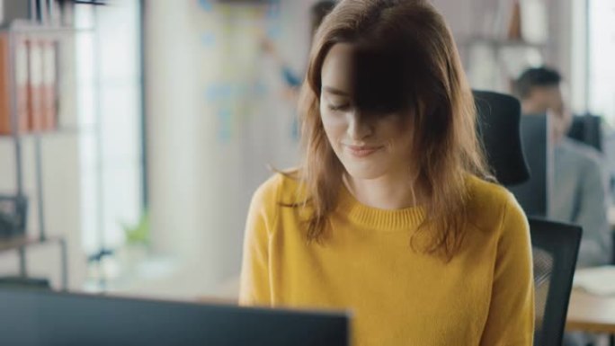 美丽聪明的红发女专家坐在办公桌前在台式电脑上工作。明亮现代的开放空间办公室，配有时尚符合人体工程学的
