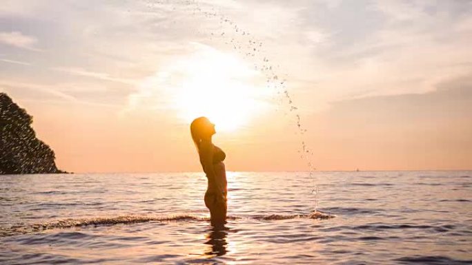 穿着比基尼的年轻女子在日落时用头发溅海水