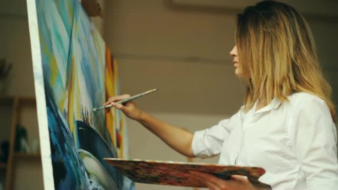 穿着白衬衫的迷人年轻女子正在用油画在帆布上画海景，拿着刷子和调色板在现代工作室工作。艺术和创造力概念