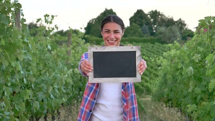 美丽的女农夫微笑着看着葡萄田，拿着一块黑色的木板，穿着衬衫，绿色的背景。