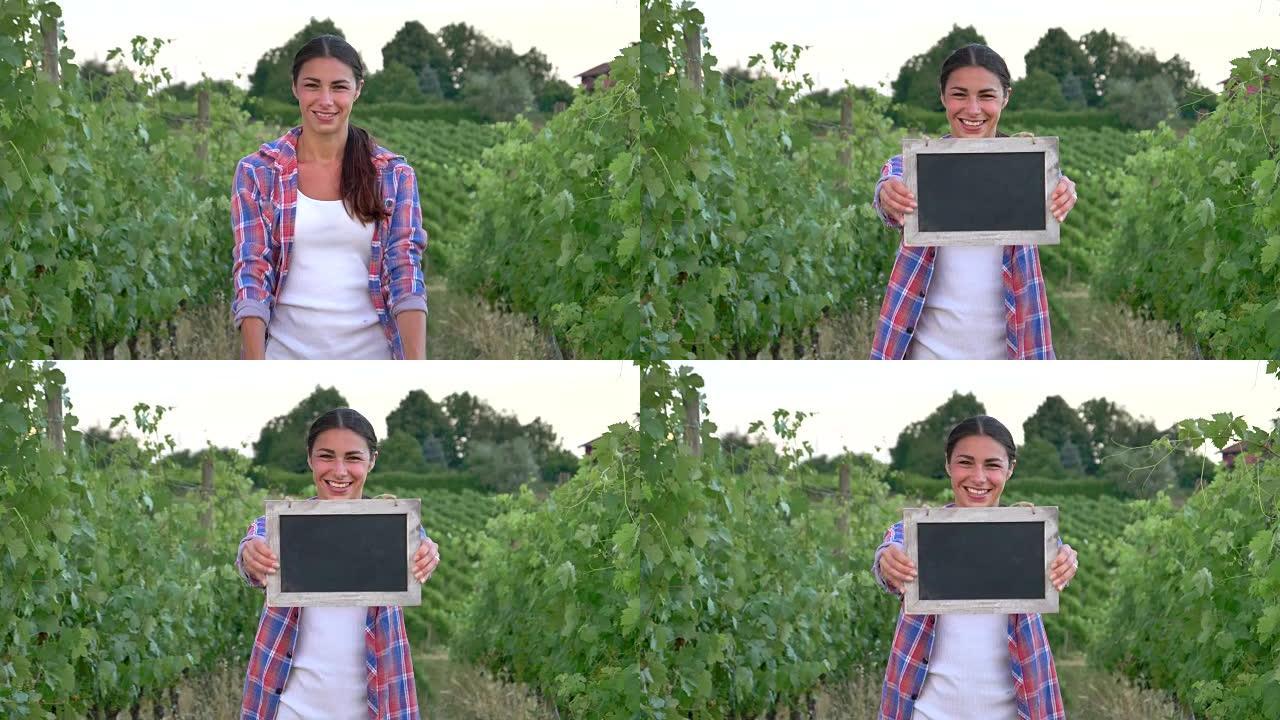 美丽的女农夫微笑着看着葡萄田，拿着一块黑色的木板，穿着衬衫，绿色的背景。