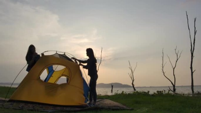年轻的亚洲女孩在夏天的湖边搭起帐篷的剪影。露营、旅行和友谊概念。