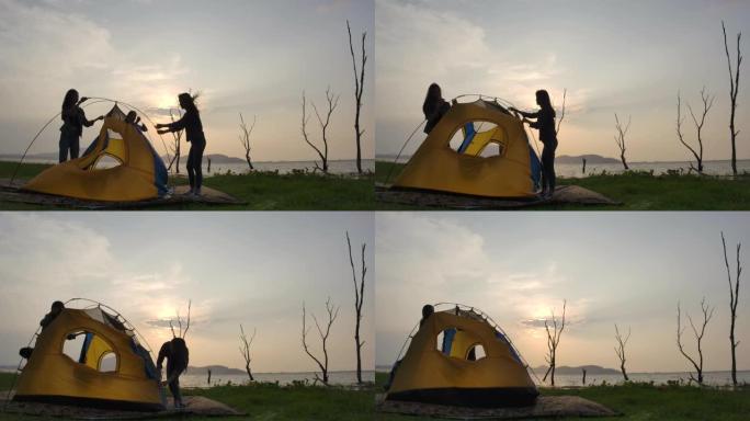 年轻的亚洲女孩在夏天的湖边搭起帐篷的剪影。露营、旅行和友谊概念。