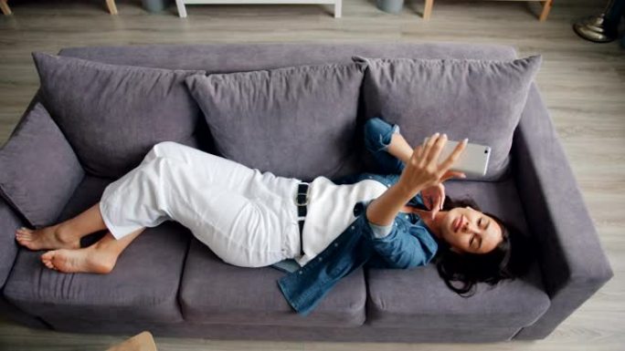 美丽的女人用智能手机相机躺在沙发上自拍的俯视图