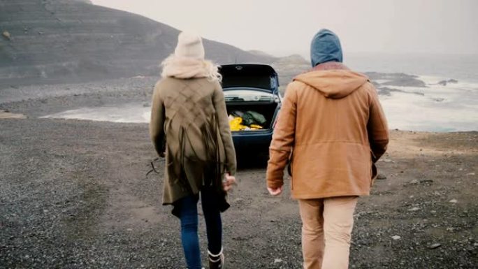 年轻幸福的夫妇开车旅行。迷人的男人和女人走到冰岛黑海滩附近的车辆上。
