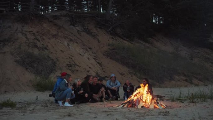 一群朋友坐在海滩上的篝火旁。浪漫的夏夜氛围。男孩和女孩喝葡萄酒和其他轻度酒精饮料。他们说话并为活着感