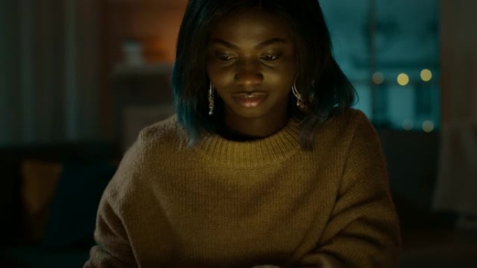 美丽的黑人女孩的肖像坐在家里的办公桌旁时使用笔记本电脑，她的脸被屏幕灯照亮。晚上，有创意的女人在舒适