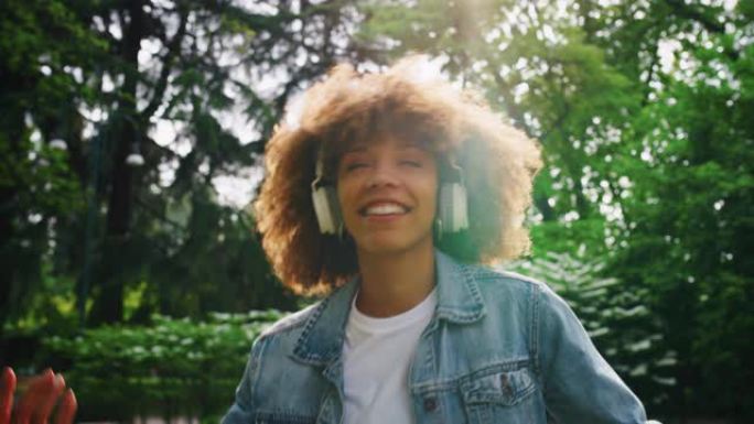 在阳光明媚的日子里，年轻的非洲妇女的慢动作很高兴戴着白色耳机听音乐并在绿色城市公园跳舞