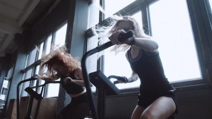 两名年轻的微笑运动欧洲妇女在有氧运动训练，慢动作时在健身房自行车器械上锻炼。
