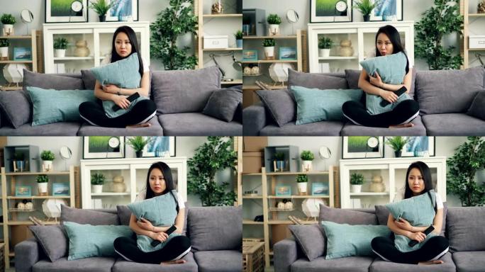 美丽的亚洲年轻女子正在家里拿着枕头在电视上看恐怖片，对可怕的情节做出反应，然后使用遥控器。电影和人的
