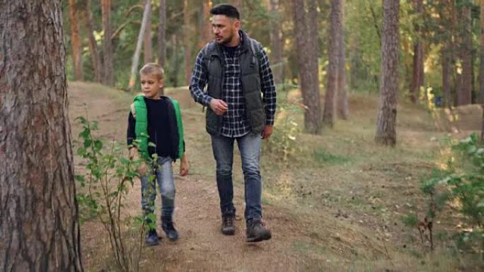 英俊的年轻人和他可爱的小儿子正在森林里散步，聊天享受新鲜的空气、自然和交流。家庭、父亲和旅游概念。