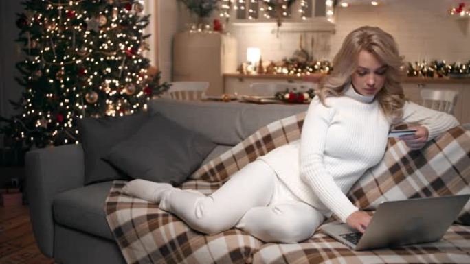 美丽的金发女郎使用坐在客厅沙发上的笔记本电脑进行圣诞节网上购物。