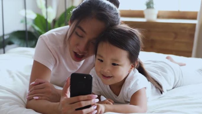 快乐的亚洲妈妈用智能手机和孩子女儿躺在床上