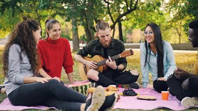 多种族的朋友团体在公园里玩得很开心，弹吉他，拍手唱歌，享受闲暇时光和温暖的秋日。人和音乐概念。