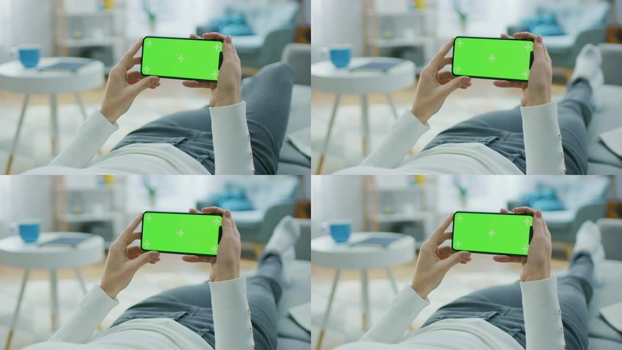 在家中的年轻女子躺在沙发上，使用水平景观模式下的绿色模拟屏幕智能手机。女孩使用手机，浏览互联网，观看