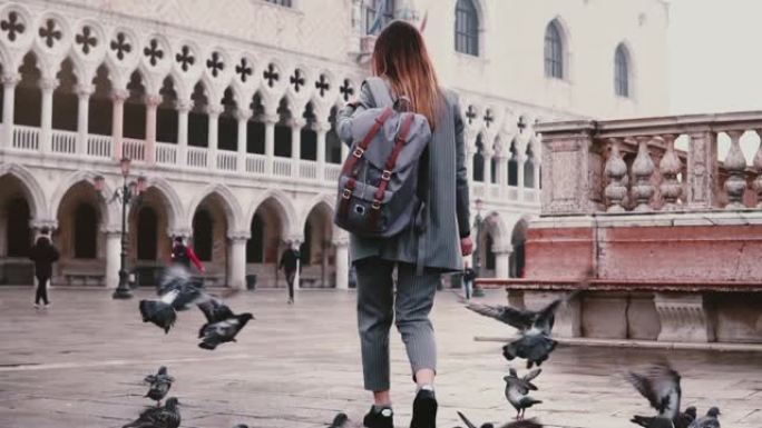 在意大利威尼斯令人惊叹的老城广场上，一大群鸽子跟随带着背包的快乐女游客慢动作