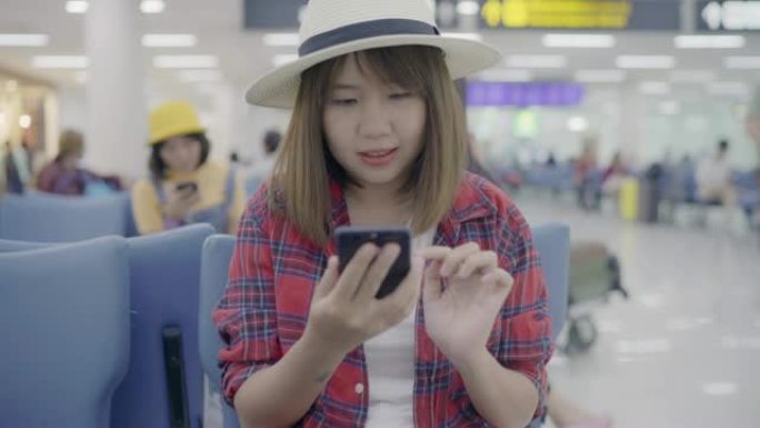 快乐的亚洲女性在国际机场登机口等待航班时，坐在候机厅的椅子上使用和检查智能手机。女人在机场概念上很开