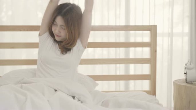 美丽迷人的年轻亚洲女子醒来，微笑着在卧室的床上伸展双臂。年轻的亚洲女性在家使用放松时间。生活方式女性