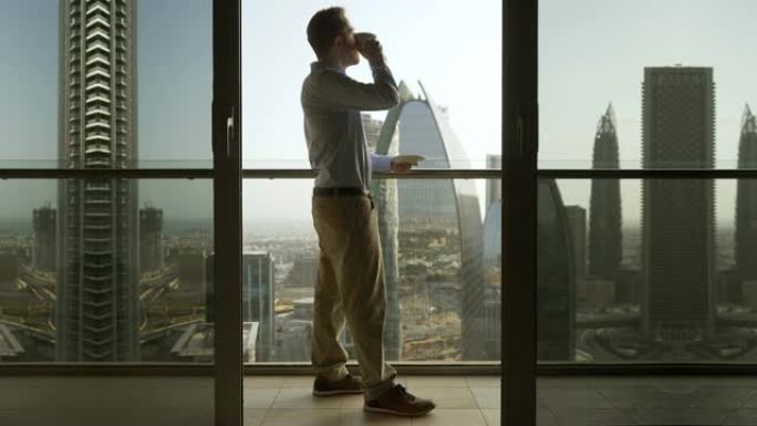 年轻的白人男子在阳台上喝咖啡，看着摩天大楼的现代城市