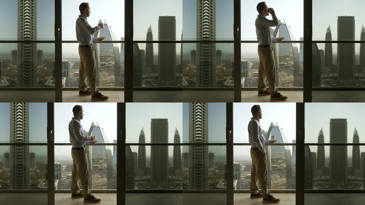 年轻的白人男子在阳台上喝咖啡，看着摩天大楼的现代城市