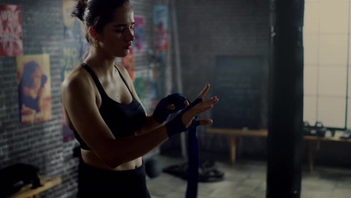 强壮而美丽的女性用手包裹着她的手，在健身房开始她的跆拳道锻炼，墙上贴着励志海报。她在手腕，拇指和指关