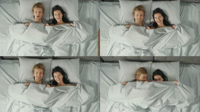 快乐有趣的年轻夫妇在床上用毯子玩躲猫猫，露出自己的脸，做出愚蠢的面孔。可爱的年轻人玩得开心。自上而下